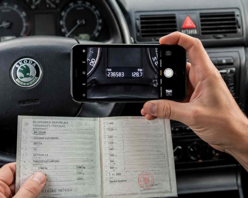 Detail ruky držiacej smartfón, ktorý fotí displej palubnej dosky staršej Škody Octavia s počtom najazdených kilometrov, v pozadí je vidieť otvorený technický preukaz – ilustrácia dôležitej fotodokumentácie pred odovzdaním auta kupujúcemu.