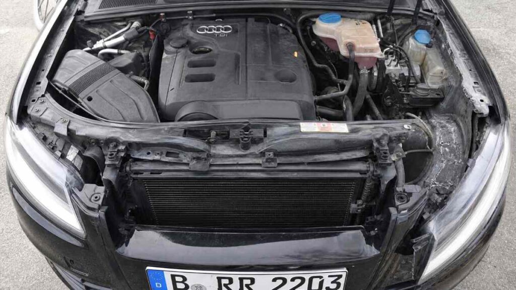 Detailný pohľad do motorového priestoru ojazdeného sedanu po dovoze z Nemecka, kde nerovnomerné zvary na pravom blatníku, čerstvý tmel okolo upevnenia chladiča a dva rozdielne odtiene čierneho laku medzi nárazníkom a kapotou jasne odhaľujú, že vozidlo prešlo závažnou opravou po čelnej kolízii, hoci predajca v inzeráte uviedol „nehavarované, prvý majiteľ, plná servisná história